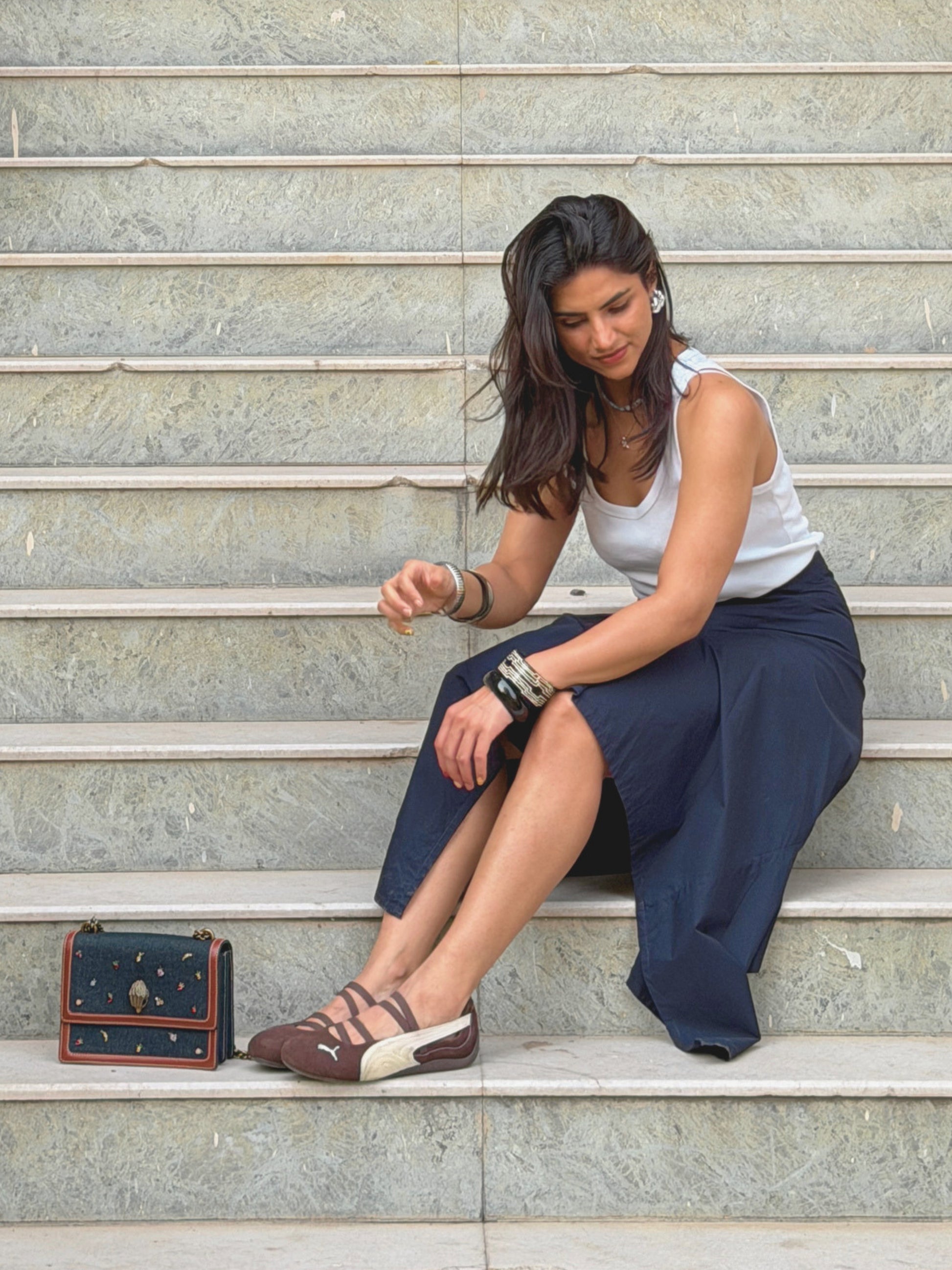 model sitting on steps wearing a white top and navy blue ikami berlin statement skirt with a small bag beside her.
