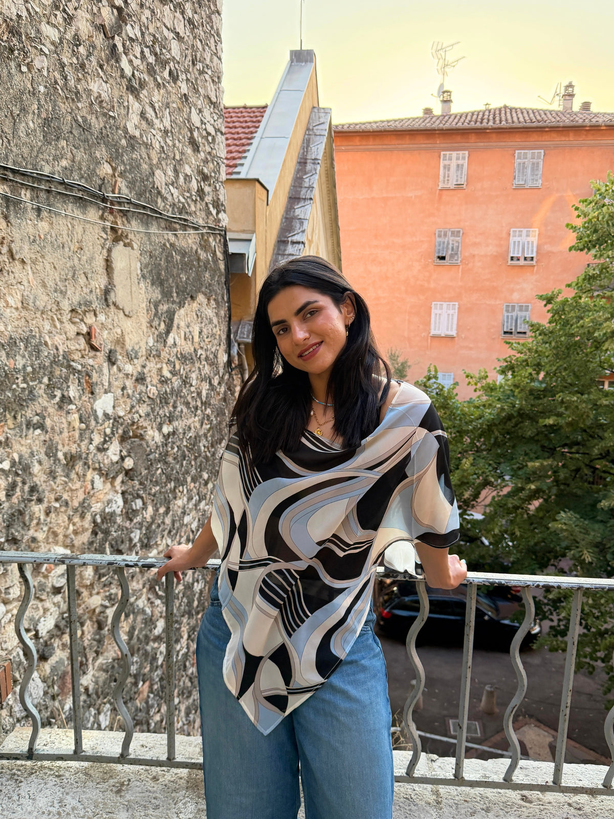 A woman wearing a black, white, and gray abstract patterned poncho and blue jeans, standing on an outdoor balcony in nice during sunset