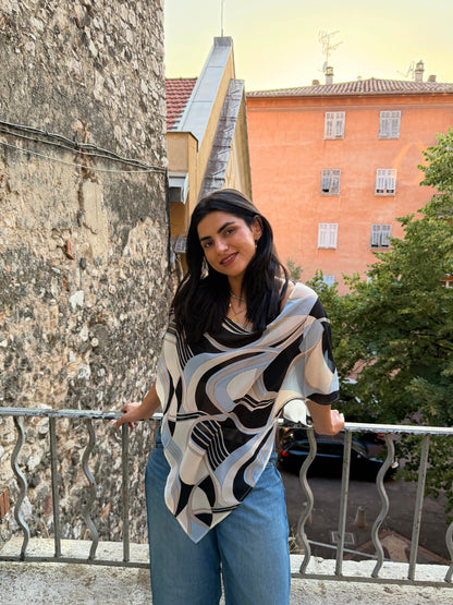 A woman wearing a black, white, and gray abstract patterned poncho and blue jeans, standing on an outdoor balcony in nice during sunset