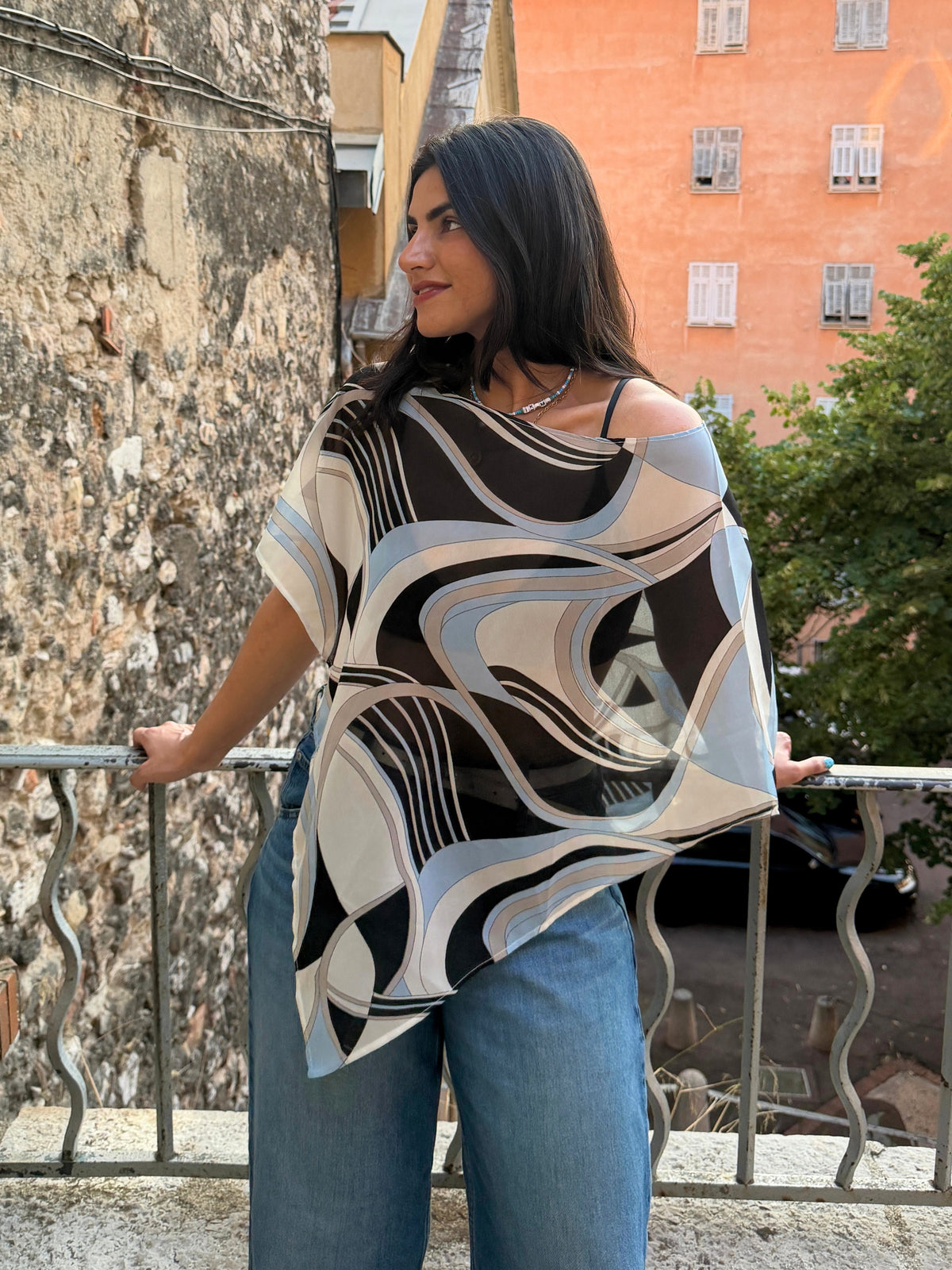 A woman wearing a loose, short-sleeve abstract-patterned top with blue jeans, standing on a balcony with a stone wall and orange building in the background in europe in ikami