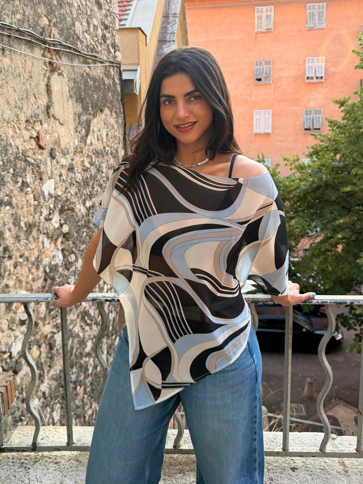 A woman wearing a loose, off-the-shoulder top with a black, white, and light blue abstract wavy pattern, paired with blue jeans, standing on a balcony with a stone wall and an orange building in the background in europe.