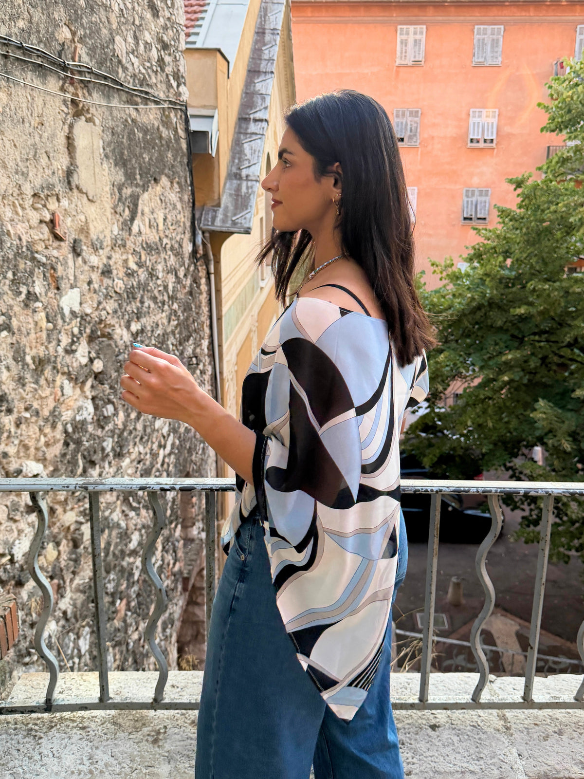 model wearing an ikami poncho which is loose, off-the-shoulder blouse with a black, white, and light blue abstract geometric pattern, paired with blue jeans, standing on a balcony in nice, france in the middle of summer.