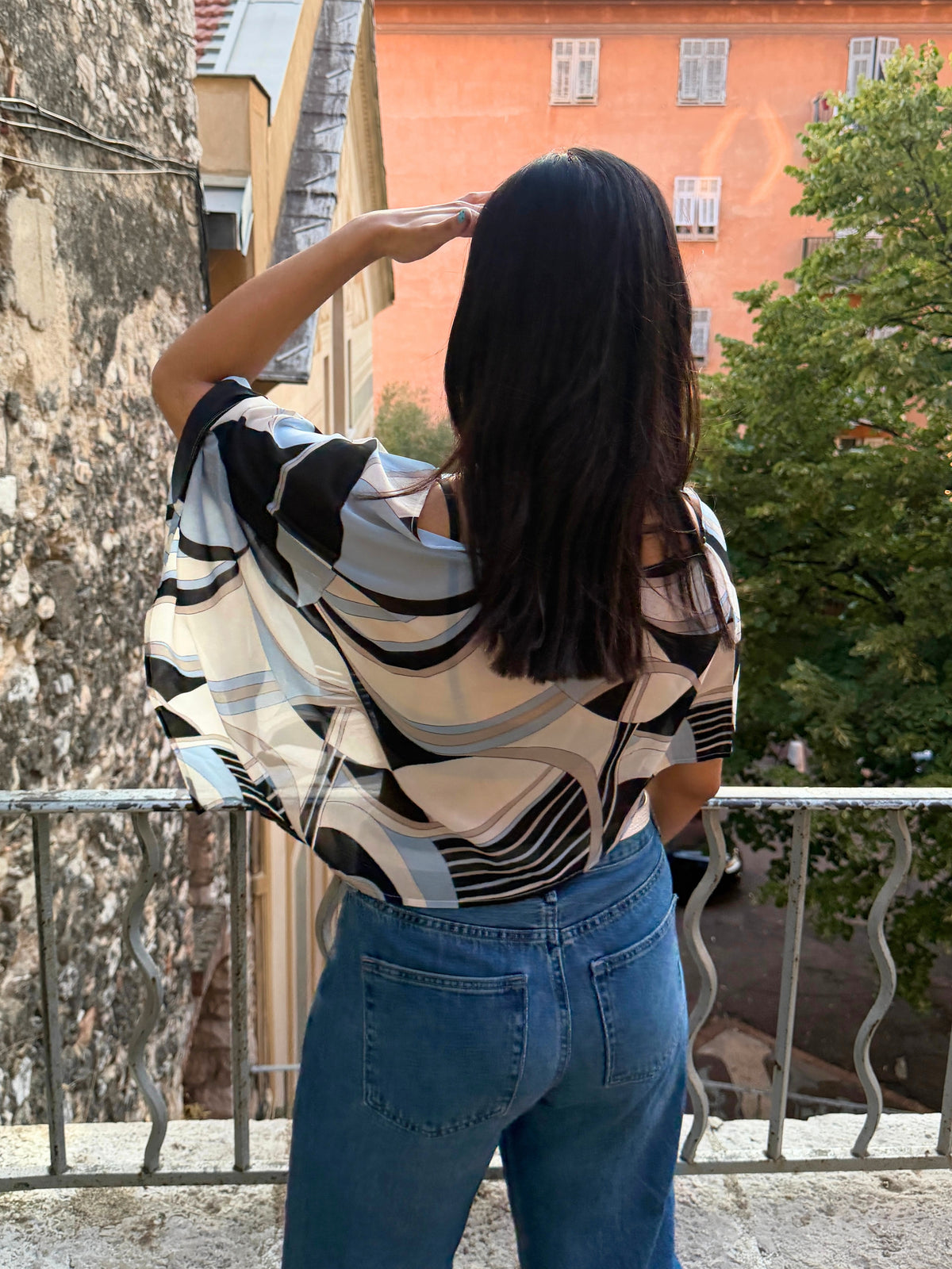 model viewed from behind wearing a loose, short-sleeve blouse with a black, white, and gray geometric pattern, paired with blue jeans, standing on a balcony with a gorgeous view